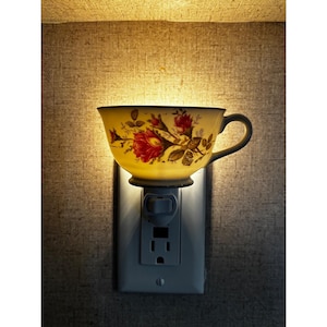 May include: A plug-in wall night light featuring a cream-colored teacup with a red rose floral pattern. The teacup is mounted onto a standard electrical outlet cover plate, emitting a warm, soft glow from within the cup.