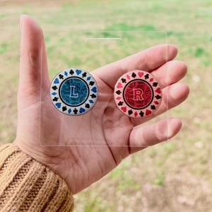 May include: Two poker chip-style buttons, one blue with a white "L" and the other red with a white "R", are held in a clear plastic holder. The buttons have suit symbols around the edges.
