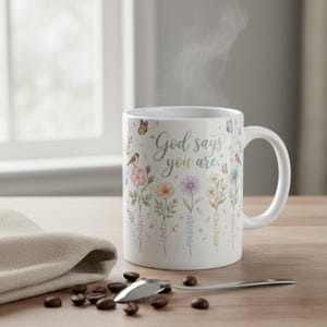May include: White ceramic mug with the text "God says you are." The mug features floral and butterfly illustrations in pastel colors. The words "Chosen, Loved, Forgiven, Redeemed, Strong" are also printed on the mug.