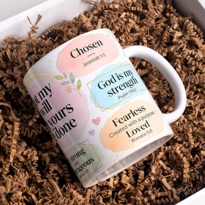 May include: White ceramic mug with inspirational quotes and floral accents. The mug features phrases like "Chosen," "God is my strength," and "Fearless." The mug is presented in a white box with brown paper filler, ideal for gifting.