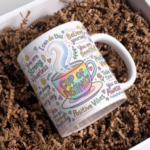 May include: White ceramic mug with a colorful "Cup of Positivity" design. The mug features inspirational phrases and a rainbow-colored cup illustration. The mug is surrounded by brown packing material.