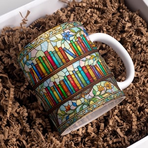 Puede incluir: Una taza de cerámica blanca con un diseño de vidriera que presenta libros coloridos en estanterías. La taza tiene un asa blanca y está colocada en una caja con relleno de papel marrón. Los libros tienen títulos como "Historia" y "Naturaleza".