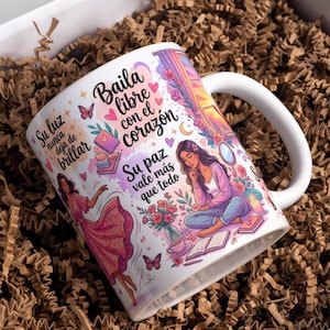 Könnte beinhalten: Weiße Keramiktasse mit einer umlaufenden Illustration mit einer Frau, Blumen, Schmetterlingen und spanischem Text. Die Tasse hat einen gebogenen Henkel und steht in einer Box mit braunem Packmaterial.