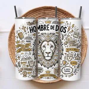 May include: Three white cylindrical tumblers with black lids and straws, featuring a detailed black and gold graphic print of a lion's face, crowns, crosses, mountains, and religious text in Spanish including "Hombre de Dios" and "Padre".
