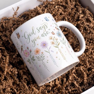 Pode incluir: Caneca de cerâmica branca com o texto "God says you are." A caneca é decorada com flores coloridas, borboletas e pássaros. As palavras "Chosen, Loved, Forgiven, Redeemed, Strong" também estão impressas.