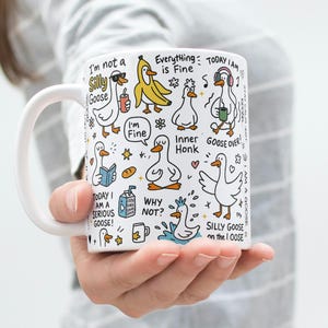 May include: White ceramic mug featuring whimsical cartoon geese with various expressions and phrases like "Silly Goose" and "Everything is Fine." The mug has a white handle and is held by a person.