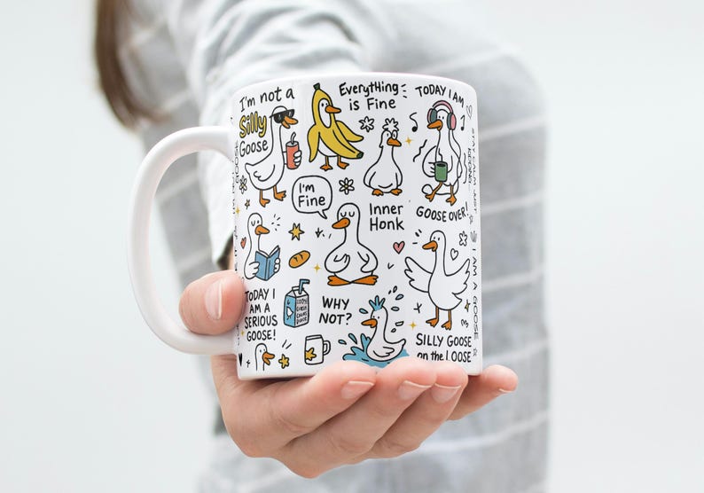 May include: White ceramic mug featuring whimsical cartoon geese with various expressions and phrases like "Silly Goose" and "Everything is Fine." The mug has a white handle and is held by a person.