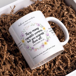 May include: White ceramic mug with a handle, featuring floral wreath and inspirational text. The text includes "Faith can move mountains" and "There many capable women, but you surpass them all." The mug is nestled in brown packing material inside a white box.