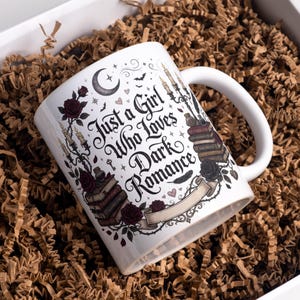 Pode incluir: Caneca de cerâmica branca com o texto "Just a Girl Who Loves Dark Romance" em escrita preta. A caneca apresenta ilustrações de rosas, velas, livros e uma lua crescente. A caneca é embalada em uma caixa branca com papel pardo.