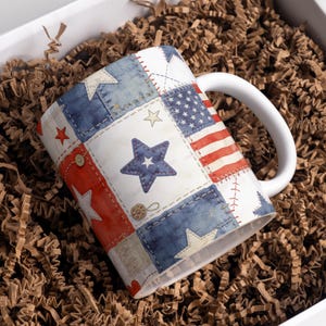 May include: White ceramic mug with a patchwork design featuring the American flag, stars, and denim-style patches in red, white, and blue. The mug has a white handle and is presented in a box with brown packing material.