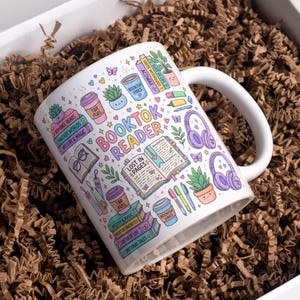 May include: White ceramic mug with a colorful "Booktok Reader" design. The mug features stacks of books, coffee cups, headphones, and the text "Lost in Pages." The design includes the text "Romance Reads," "Fantasy Worlds," and "Mystery & Magic."
