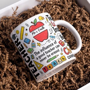 Puede incluir: Taza de cerámica blanca con asa, decorada con ilustraciones escolares y el texto "TEACHER". Presenta un diseño de manzana roja con el nombre "Mr & Smith". La taza está llena de material de embalaje marrón dentro de una caja blanca.