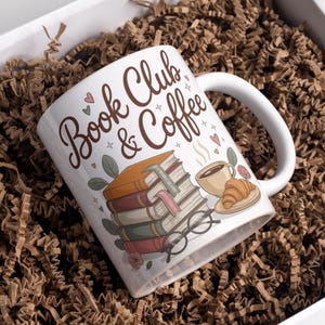 Op de afbeelding: Witte keramische mok met de woorden "Book Club & Coffee" in bruin script. De mok heeft illustraties van opgestapelde boeken, een kopje koffie, een croissant en een bril. De mok ligt in bruin verpakkingsmateriaal.