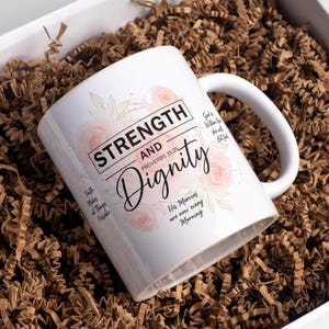 May include: White ceramic mug with the words "Strength and Dignity" and floral accents. The mug also features the text "Proverbs 31:25" and other inspirational phrases. The mug is packaged in a white box with brown paper filler.