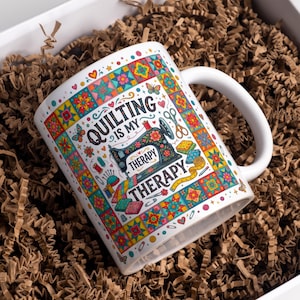 Può includere: Tazza in ceramica bianca con la scritta "Quilting is my Therapy" e un'illustrazione di una macchina da cucire. La tazza presenta un colorato design patchwork con farfalle, fiori e strumenti di cucito. La tazza è all'interno di una scatola bianca con materiale di imballaggio marrone.