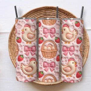 May include: Three pink insulated tumblers with black straws, featuring a repeating pattern of chickens, strawberries, bows, and Easter baskets. The tumblers are displayed on a woven basket.