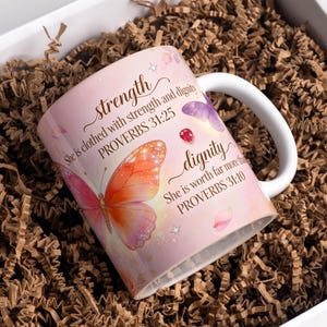 May include: A pink ceramic mug with a white handle, featuring butterfly illustrations and the words "strength" and "dignity" with Bible verses. The mug is packaged in a white box with brown paper filler.