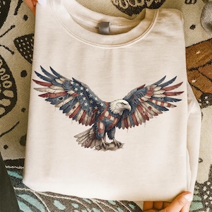 May include: A cream-colored sweatshirt featuring a detailed graphic of an American bald eagle with outstretched wings. The eagle's wings and body are adorned with a patriotic design incorporating the American flag's stars and stripes. The sweatshirt has a crew neck.