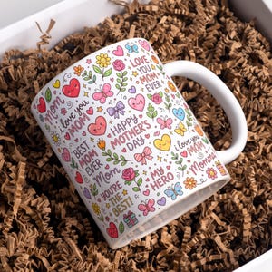Op de afbeelding: Witte keramische mok met een handvat, versierd met kleurrijke harten, bloemen, vlinders en de tekst "Happy Mother's Day". De mok bevat zinnen als "I love you Mom" en "Best Mom Ever".