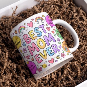 Op de afbeelding: Witte keramische mok met de woorden "BEST MOM EVER" in kleurrijke bubbelletters, versierd met regenbogen, harten, bloemen en tekstballonnen. De mok ligt in bruin verpakkingsmateriaal in een witte doos.