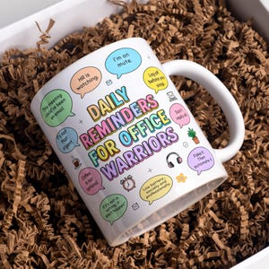 May include: White ceramic mug with the text "DAILY REMINDERS FOR OFFICE WARRIORS" in colorful bubble letters. The mug has various office-related reminders in speech bubbles, such as "I'm on mute" and "Coffee is not optional."