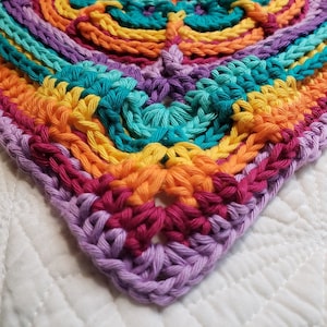 Pode incluir: Um quadrado de crochê colorido com um padrão geométrico em tons de roxo, turquesa, laranja e amarelo.