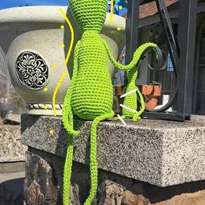 Puede incluir: Una muñeca de amigurumi de color verde lima hecha a crochet con brazos y piernas largos sentada en un muro de piedra. La muñeca tiene una cara simple con dos ojos negros.