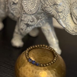 May include: A beaded bracelet with gold-colored beads and a few blue beads is displayed on a gold-colored, round object. In the background, a silver elephant statue with intricate patterns is visible.