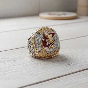 Könnte beinhalten: Ein gold-silberner Meisterschaftsring mit einer großen, runden Oberfläche. Die Oberfläche zeigt ein rot-goldenes Logo mit dem Wort "CHAMPIONS" in goldfarbenen Buchstaben. Der Ring ist mit kleinen, klaren Steinen besetzt.