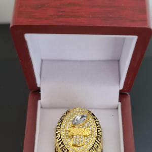 Puede incluir: Un anillo de campeonato dorado con una gema grande y transparente en el centro, rodeada de piedras más pequeñas. El anillo está en una caja de madera roja con interior blanco. El año "2026" es visible en el anillo.