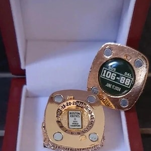 Puede incluir: Un anillo de campeonato dorado con el texto "BOSTON CELTICS 2034 WORLD CHAMPIONS" y el marcador "106-88" sobre un fondo verde. El anillo se muestra en una caja roja con interior blanco.