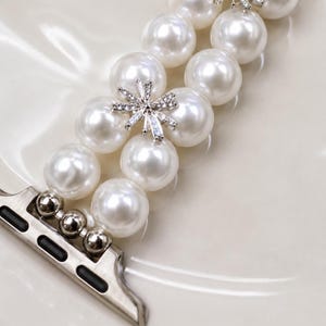 May include: White pearl watch band with silver-tone hardware. The band features large, round pearls and sparkling rhinestone accents in a floral design. The watch band is designed to attach to a smartwatch.