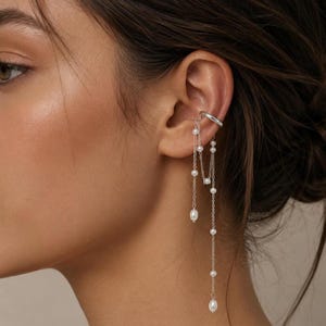 May include: A silver ear cuff featuring two delicate chains that drape downward. The chains are adorned with small, white, oval-shaped pearls spaced at intervals, with larger pearl drops at the ends of the chains, creating a layered effect.