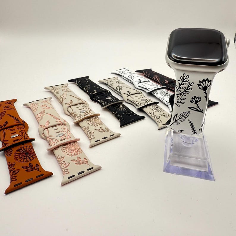 May include: A collection of silicone watch bands featuring various floral line art patterns in colors including burnt orange, cream, black, and white. One white band is attached to a smartwatch on a clear stand, while others are laid out.