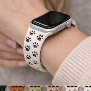 May include: A silver smartwatch with a white band featuring a pattern of black paw prints. The watch is on a wrist, and the band is secured. The image also shows a variety of colored watch bands.