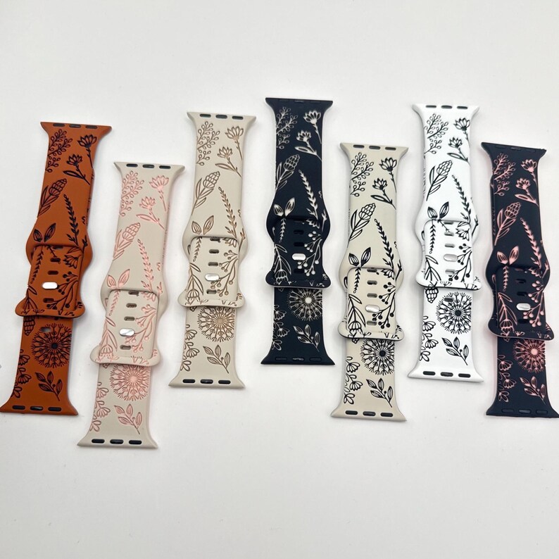May include: Seven silicone watch bands arranged in a row, featuring various floral and botanical line art patterns. The colors include burnt orange, cream, black, and white, with each band displaying unique wildflower illustrations and multiple adjustment holes.