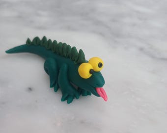 May include: A whimsical, handmade teal lizard figurine with large yellow eyes and a pink tongue. The lizard has a spiky ridge down its back and is posed on a white marble surface. This playful sculpture is a unique decorative item.