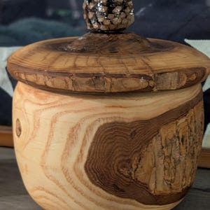 May include: A handcrafted wooden jar with a lid. The jar is made of light and dark brown wood, with visible grain patterns. The lid is round, and the knob is a small, dark brown pinecone. The jar is likely used for storage.