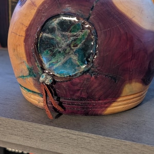 May include: A handcrafted wooden bowl with a circular, embedded, green and blue stone design. The bowl is made of wood with a mix of reddish-brown and light brown tones. A small, decorative tassel hangs from the bowl.