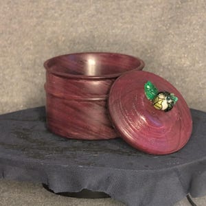 May include: A round, burgundy-colored wooden box with a lid. The lid is slightly ajar, revealing the interior. The box has a decorative, swirling grain pattern. The lid is adorned with a small, colorful, decorative element.