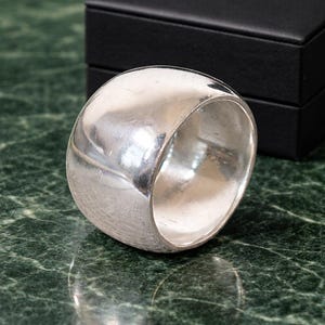 May include: A broad, silver ring with a rounded, smooth surface. The ring is highly reflective, catching light and creating highlights. The ring is set against a dark background, and the ring's design is simple and modern.