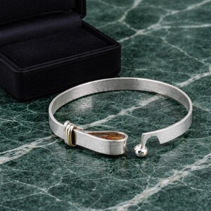 May include: A silver-colored bracelet with a unique clasp design. The bracelet has a hammered texture and a teardrop-shaped clasp with a gold accent. The open end features a small silver ball. The bracelet sits on a green marble surface.