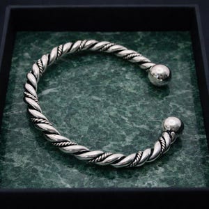 May include: A silver-colored, twisted-rope design cuff bracelet with a ball at each end. The bracelet is displayed on a green marble-like surface inside a black box. The bracelet's design is a spiral of interwoven strands.