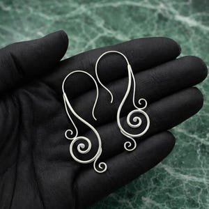 Puede incluir: Un par de pendientes plateados con diseño de espirales abstractas. Cada pendiente presenta un gancho largo y curvado que conduce a un diseño fluido y abierto con una espiral en la parte inferior. Los pendientes se muestran sobre un fondo negro.