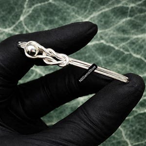 Könnte beinhalten: Eine silberfarbene Krawattenklammer mit Knoten-Design und kleinem Kugelakzent. Die Klammer wird von einer behandschuhten Hand gehalten. Der Text "NDGemsStore" ist auf der Klammer sichtbar.
