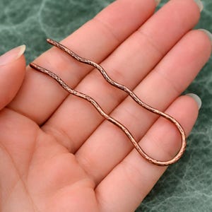 May include: A copper-colored hair pin with a hammered texture. The pin is held in a hand against a green background. The pin has a wavy design.