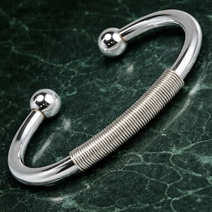 May include: A silver-toned cuff bracelet with a curved design. The bracelet features two spherical end caps and a central section wrapped in coiled wire. The background is a dark green, marbled surface.