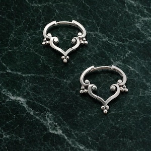 May include: Two silver-colored hoop earrings with a heart-shaped design. Each earring features a series of small, round embellishments and a decorative, curved design. The earrings are displayed on a dark green marble surface.