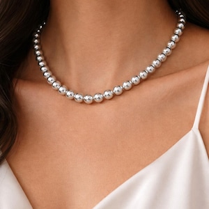 May include: A silver beaded necklace with a graduated design. The necklace features multiple round, shiny beads that vary in size, creating a stylish and elegant look. The necklace is worn against a white top.