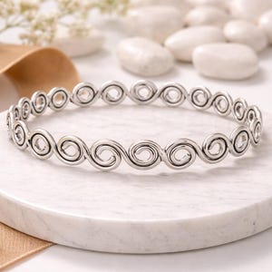 May include: A silver-colored bangle bracelet with a continuous swirl design, resting on a white marble surface. The bracelet is the focal point, with white pebbles and floral accents in the background.